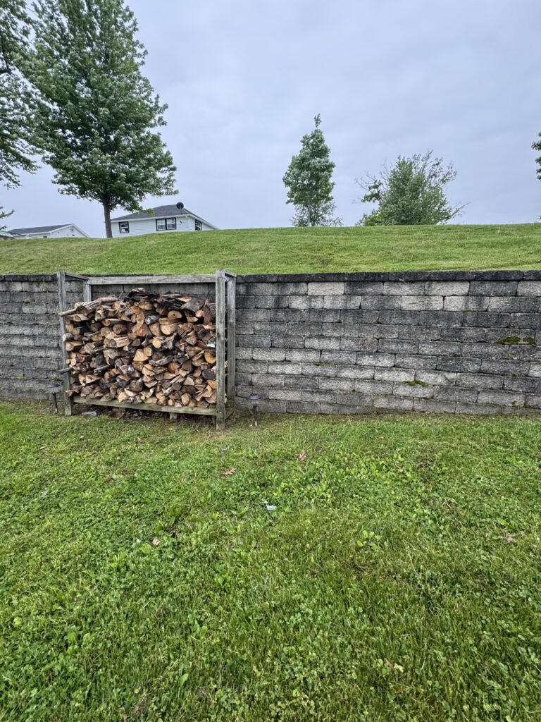 Woodpile stacked on a wooden rack beside a short stone wall, with a green lawn and houses in the background on a cloudy day.