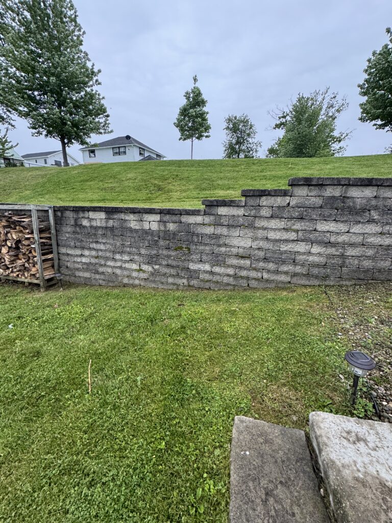 Green lawn with a stacked stone wall and woodpile on the left, and concrete steps on the right, with a small house in the distance.