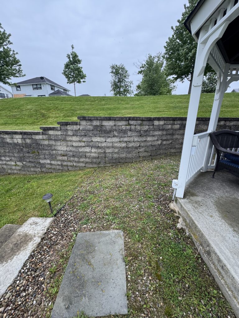Backyard scene showing a layered stone retaining wall, gravel path, and stepping stones leading to a grassy hill with a white porch railing on the right and trees in the distance under a cloudy sky.