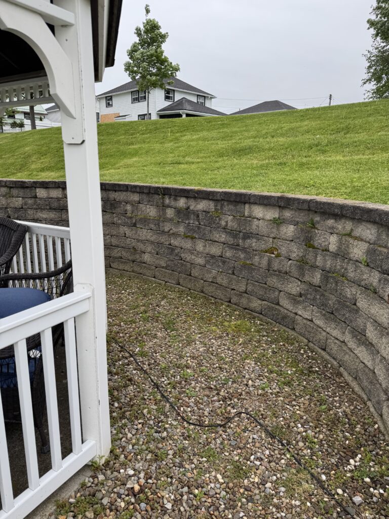 Gravel patio with a white railing on the left, a stacked-stone retaining wall, and a grassy hill rising toward white houses under a cloudy sky.