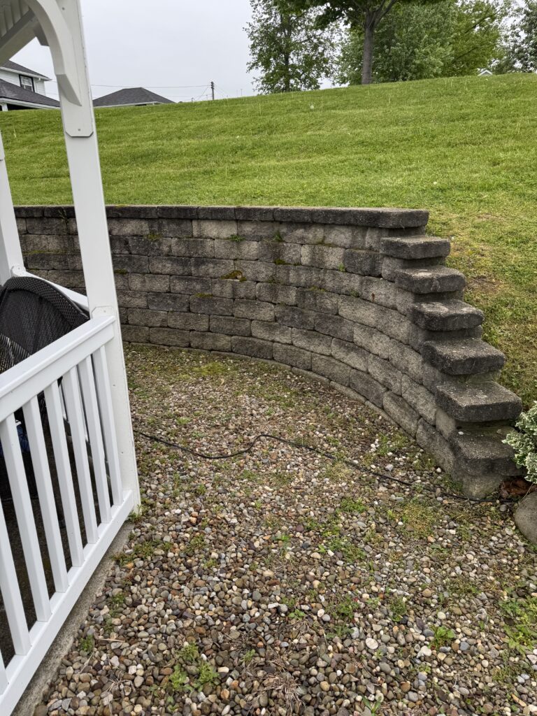 Stone block retaining wall with a narrow stair-step leading up, seen from a gravel-covered ground near a white porch railing and grassy hill in the background.