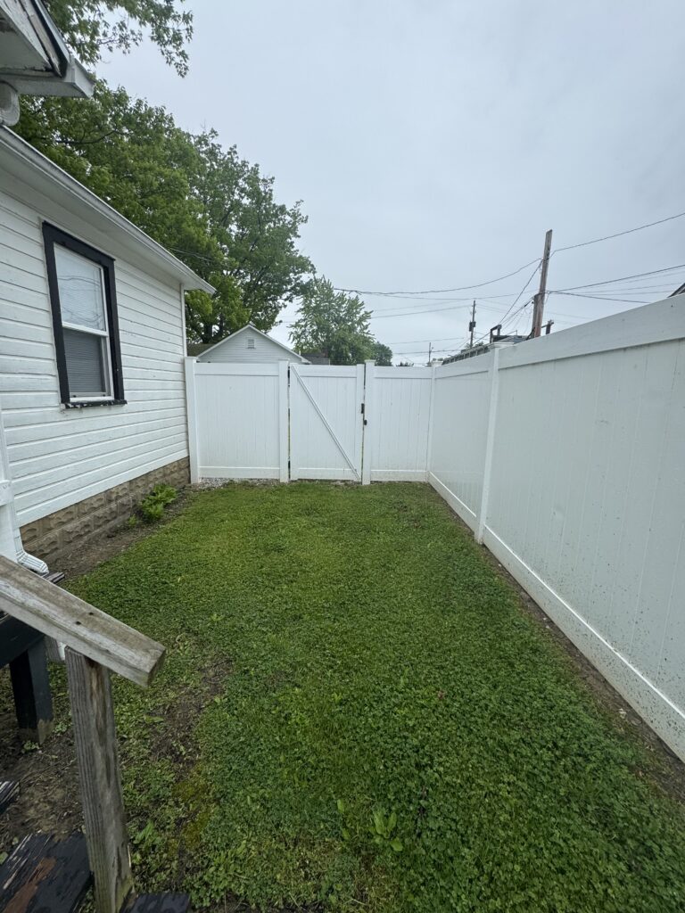 Backyard scene with a white fence and central gate, green lawn, and the side of a white house with a window to the left, under a cloudy sky.