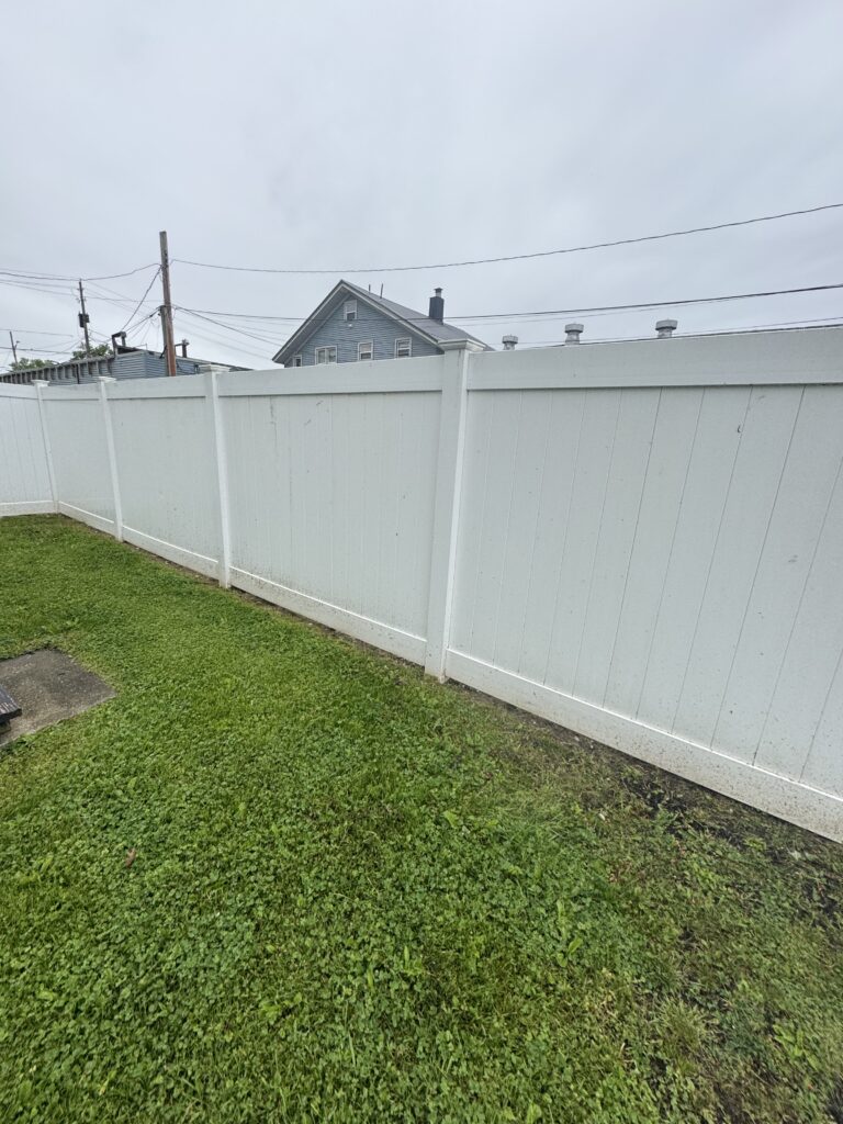 White privacy fence over a green lawn with a blue, pitched-roof house in the background under a cloudy sky.