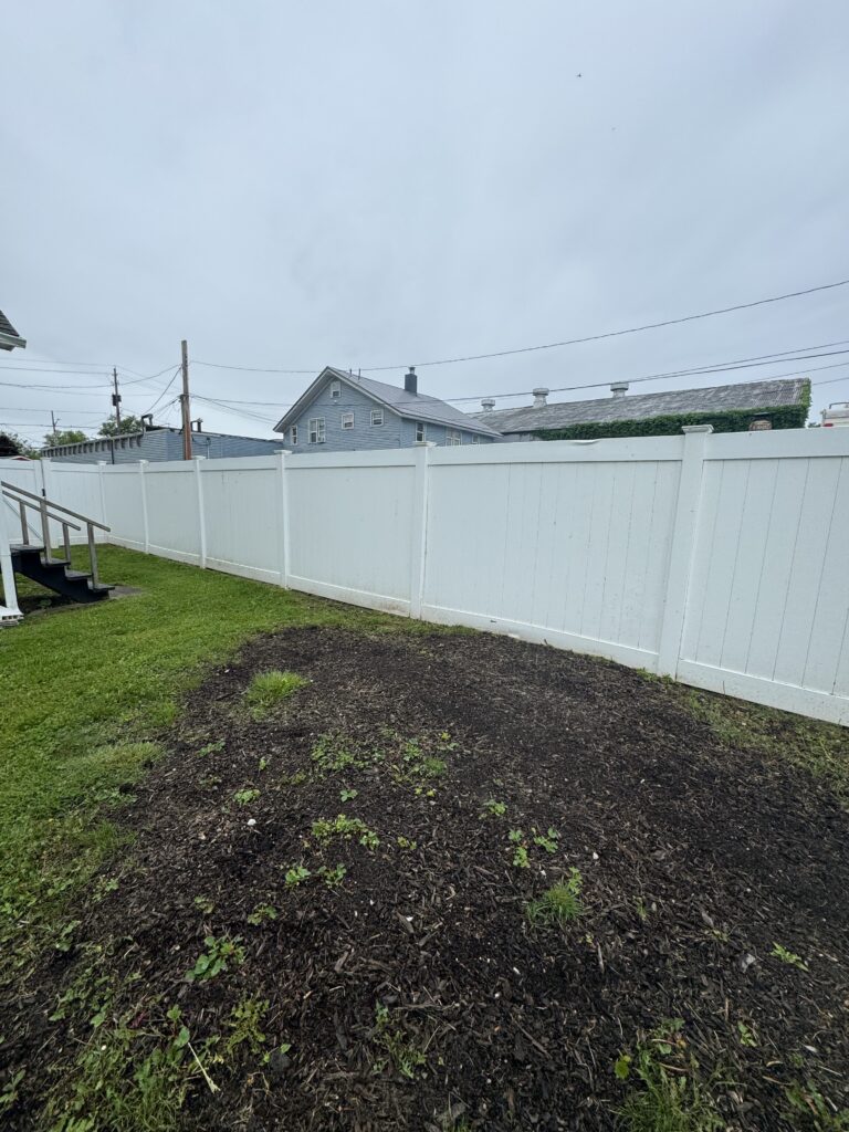 Backyard scene with a white vinyl fence, mulched bed, and a blue house in the background under a cloudy sky.