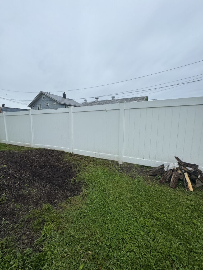 Backyard scene with a tall white fence, blue house in the background, and a pile of split logs on the right under a cloudy sky nearby.