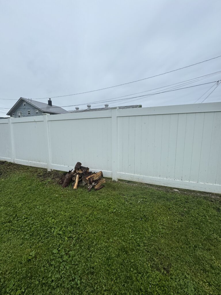 White privacy fence along a grassy yard with a small woodpile in the foreground and utility lines across a pale blue sky, house peeking from left edge