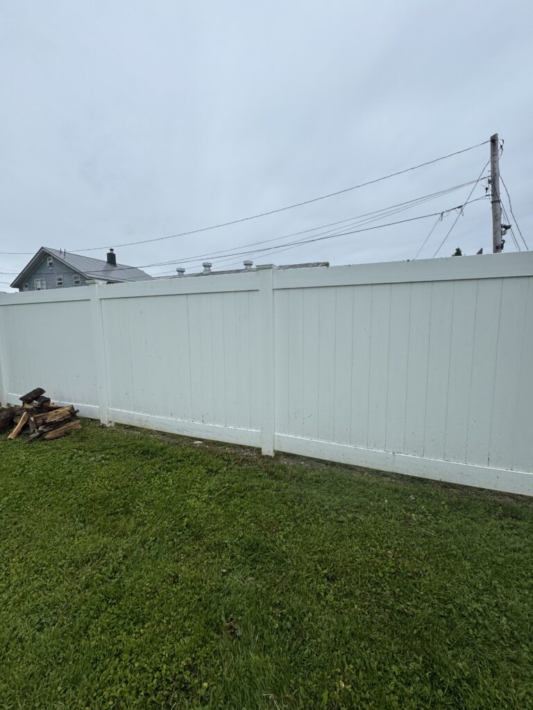 White wooden fence runs across a grassy backyard with a suburban house and utility pole in the distance; a small pile of firewood sits near the fence on the left.