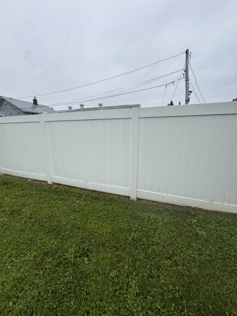 White privacy fence in the foreground, with green grass and a utility pole with power lines in the distance under a cloudy sky.