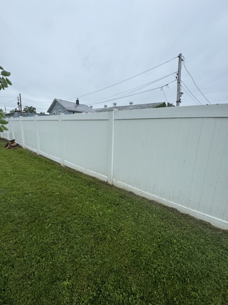 Backyard scene with a tall white privacy fence, green lawn, and utility poles with power lines under a cloudy sky.
