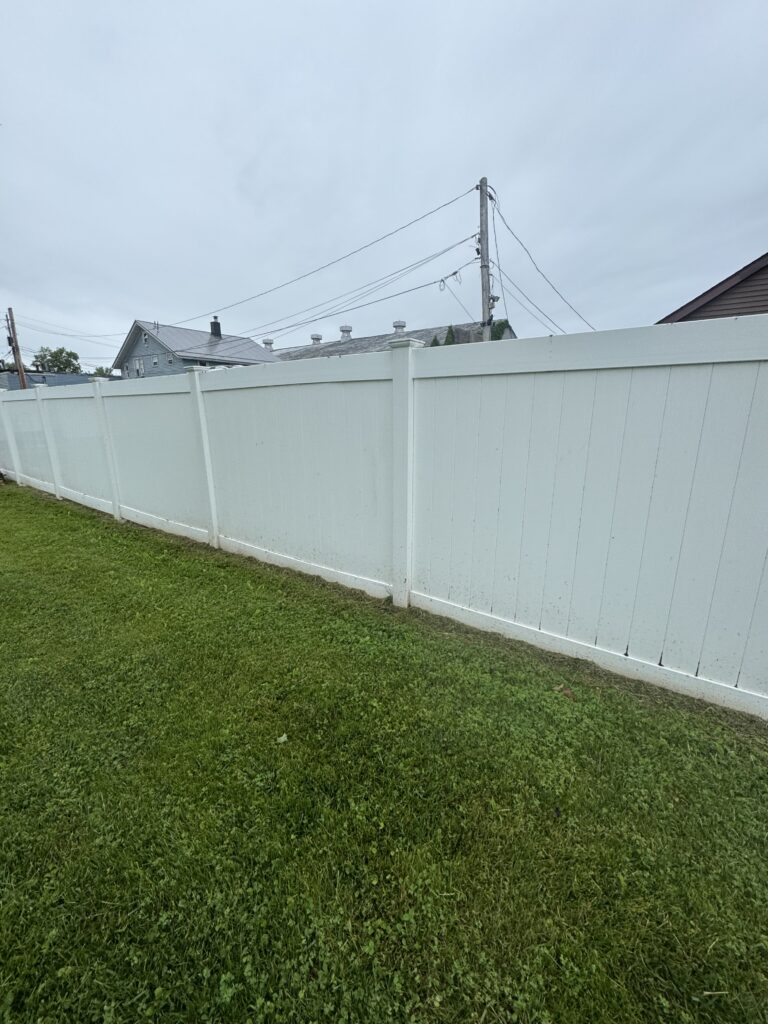 White vinyl backyard fence runs along a green lawn with suburban houses and a power pole in the distance under a gray sky.
