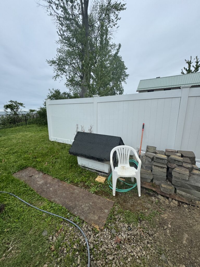Backyard scene with a white privacy fence, a large tree, and a white plastic chair near a stack of bricks and a coiled hose on a gravel and grass surface.