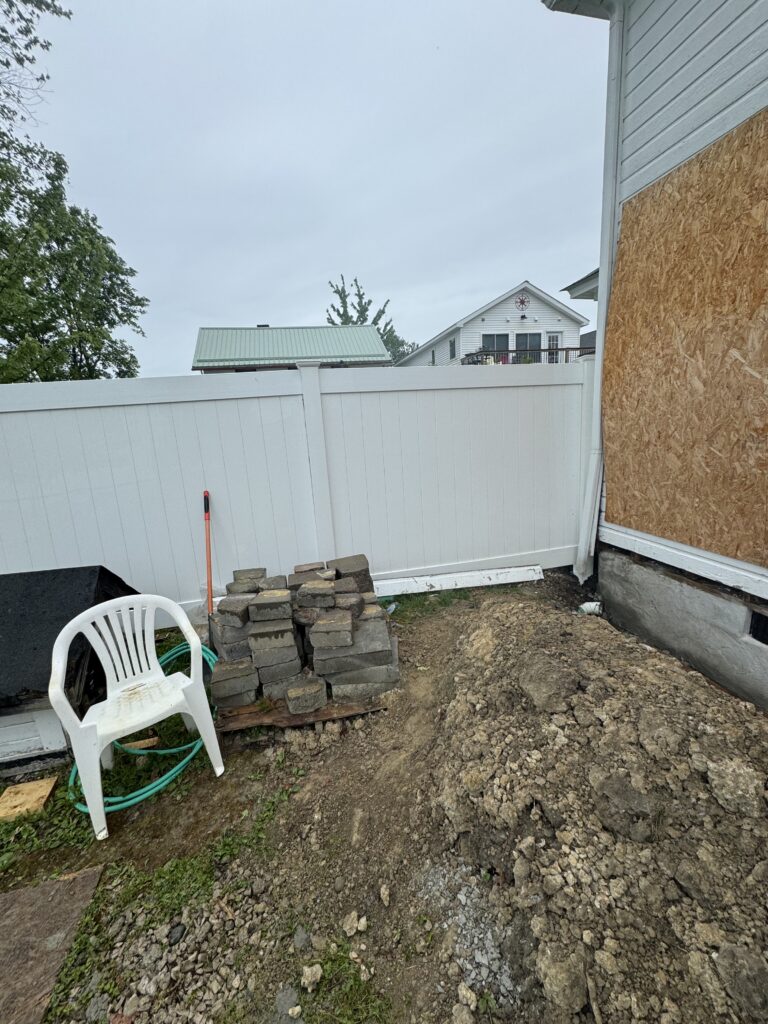 Backyard construction area with dirt ground, a white plastic chair, hose, and a stack of bricks next to a plywood-covered wall.