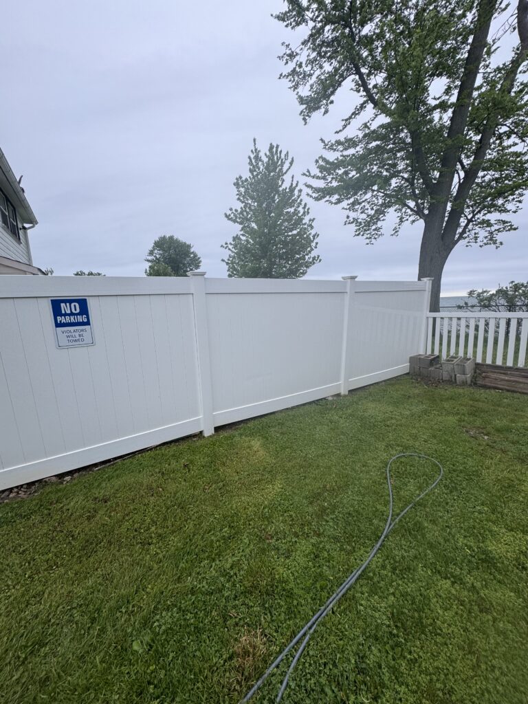 Backyard scene with a tall white fence, green lawn, and a coiled hose along the ground; a blue No Parking sign is mounted on the fence.