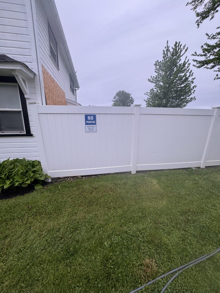 White backyard fence with a No Parking sign, a two-story white house, boarded window area, and green lawn in the foreground.