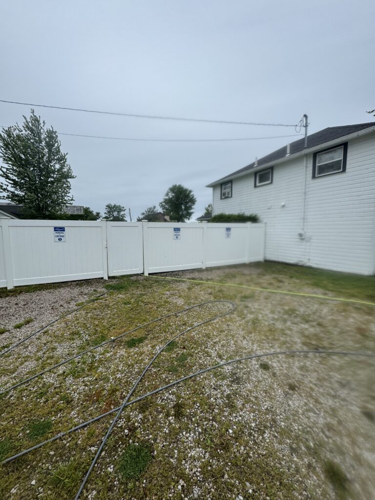 Backyard scene with a white privacy fence, a two-story white house on the right, and hoses lying on gravel and grass in the yard.