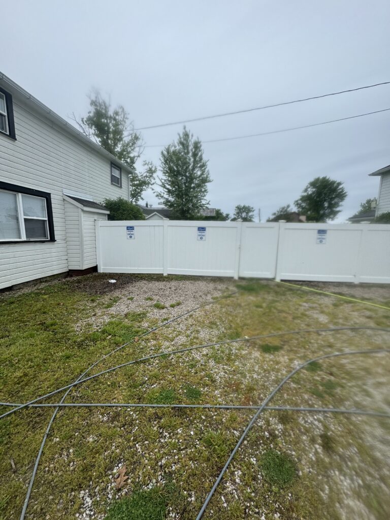 Backyard scene with a white house on the left, a tall white fence, and garden hoses sprawled across a gravel and grass yard under an overcast sky.