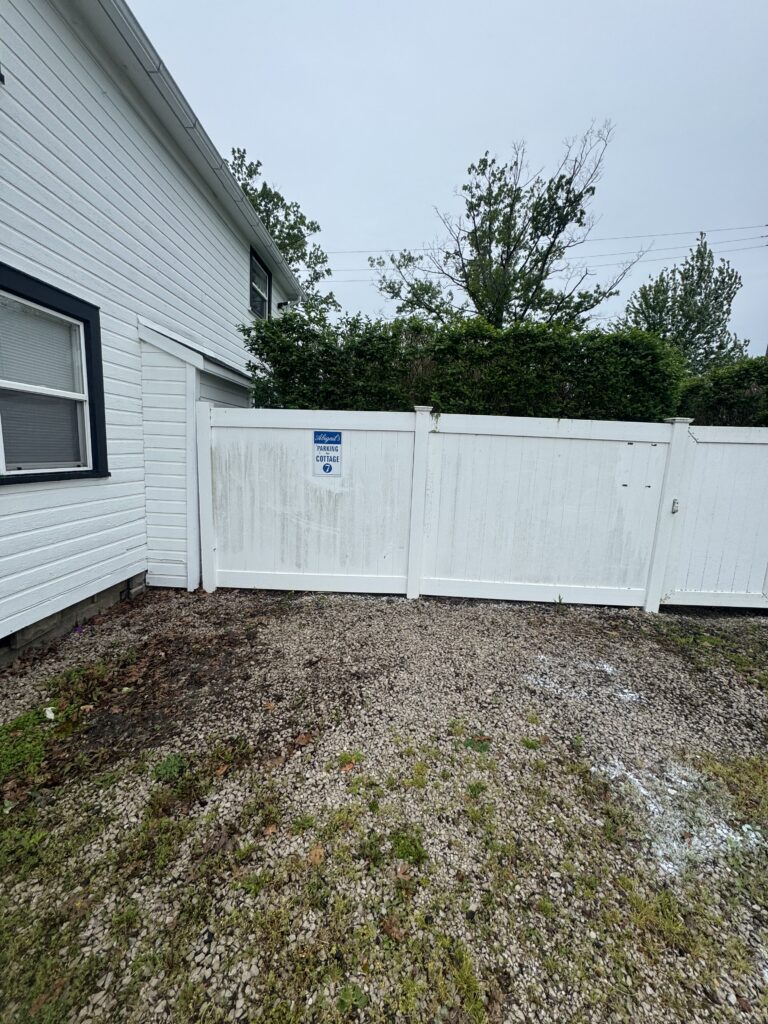 White wooden fence with a blue sign in front of a gravel yard beside a white house; overcast sky and trees beyond the fence.