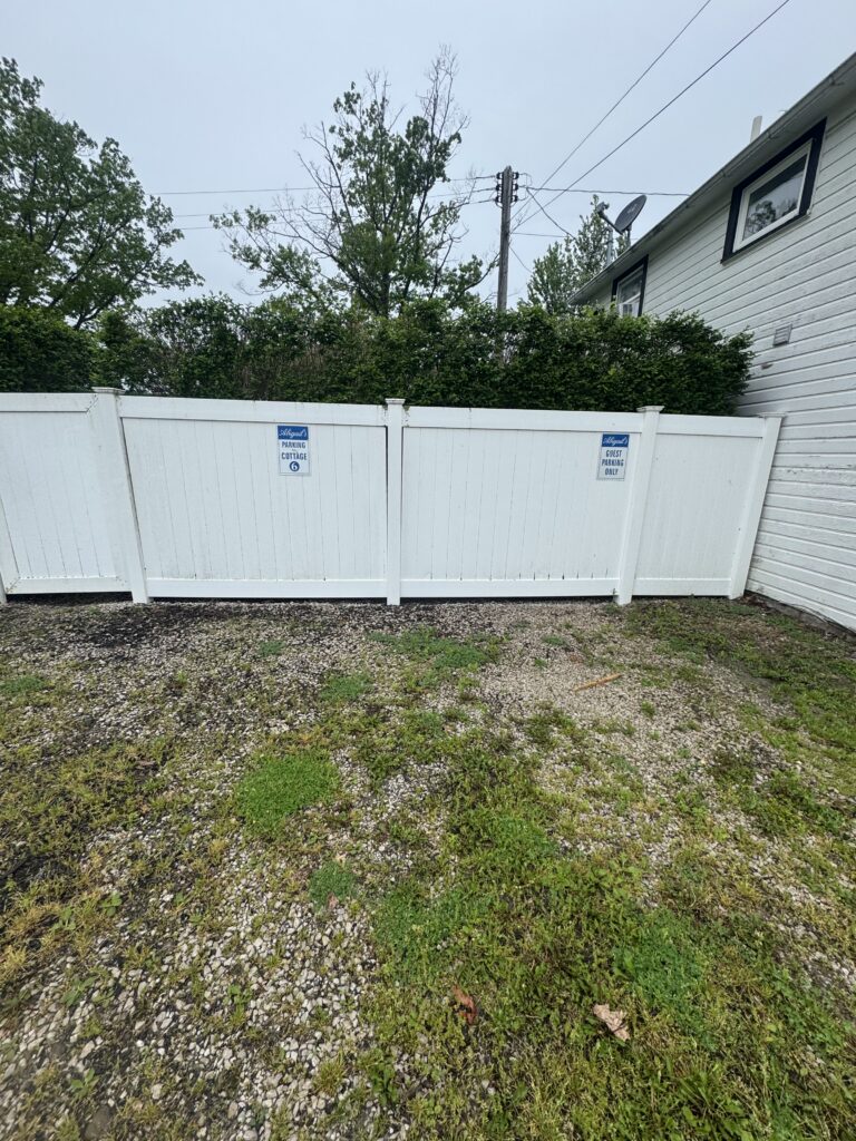 White privacy fence with blue parking signs, backed by hedges and a house with a satellite dish.