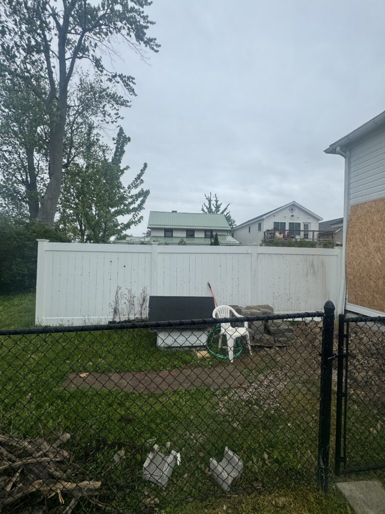 Backyard scene with a white privacy fence, chain-link fence in the foreground, a white plastic chair, hose, and scattered debris under an cloudy sky.