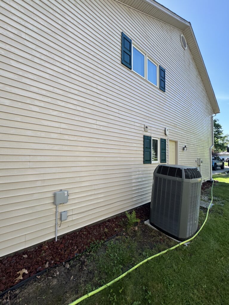 Side of a beige house with blue shutters over a two-pane window; large outdoor air conditioner unit and a yellow hose in the foreground.