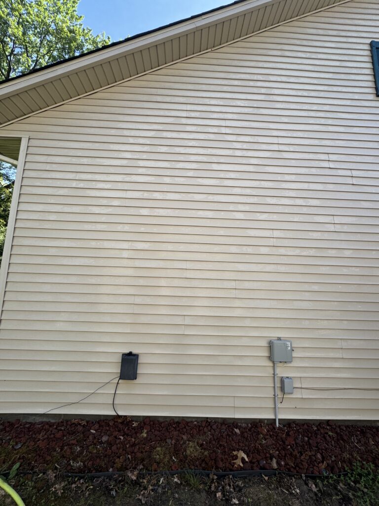 Exterior view of a beige vinyl-sided house wall with a slanted roof edge and visible utility boxes near the ground; a mulch bed runs along the base.