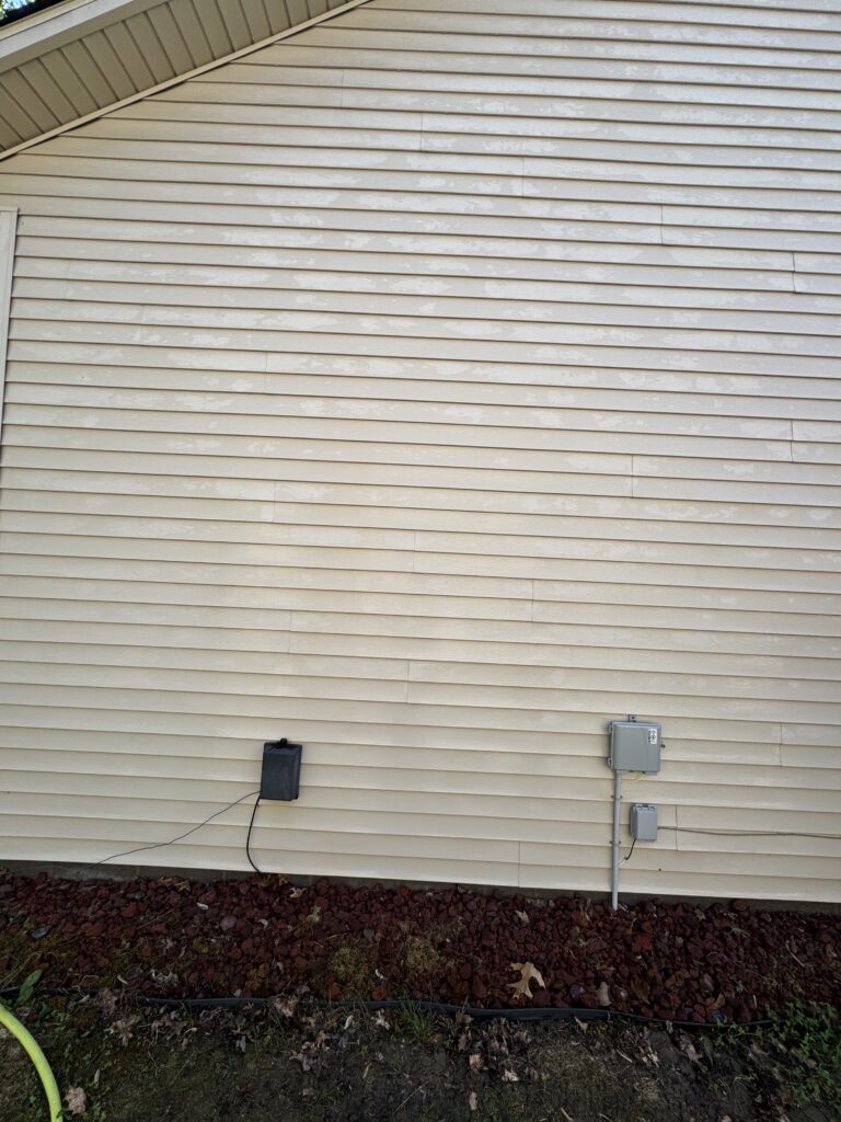 Exterior beige house siding with utility meters and electrical box mounted on the wall; a green garden hose lies along the ground nearby.