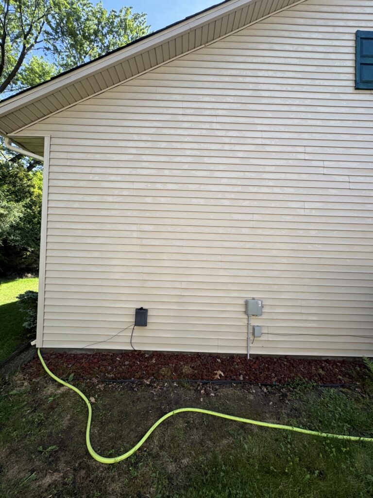 Side of a beige house with horizontal siding, yellow garden hose snaking along the ground, and electrical meters near the base yard.
