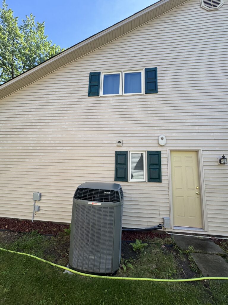 Front of a beige house with blue shutters, a double window, and a pale yellow door; an outdoor AC unit sits in the yard nearby.