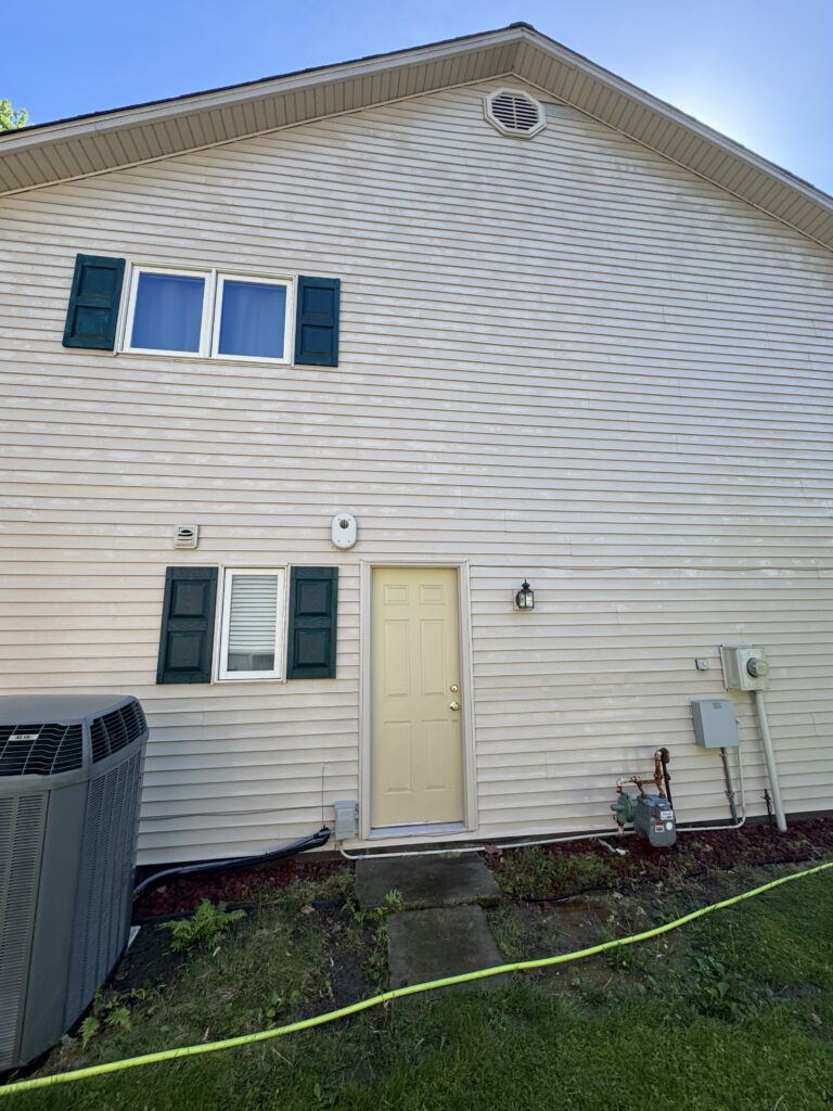 Side of a beige house with green window shutters, a pale yellow door, and outdoor utilities (AC unit, gas meter); a yellow hose runs across the yard.