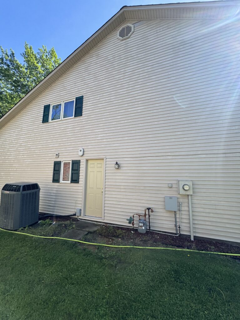 Back of a beige siding house with a cream door, green shutters, and a small window; utility meters and an air conditioner visible on the wall; green lawn foreground.