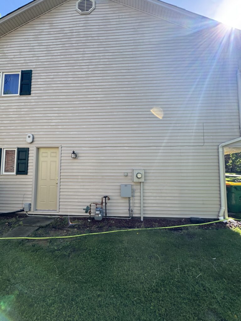 Back of beige siding house wall with a door, window with green shutters, and utility meters; sun glare creates lens flare across the image.