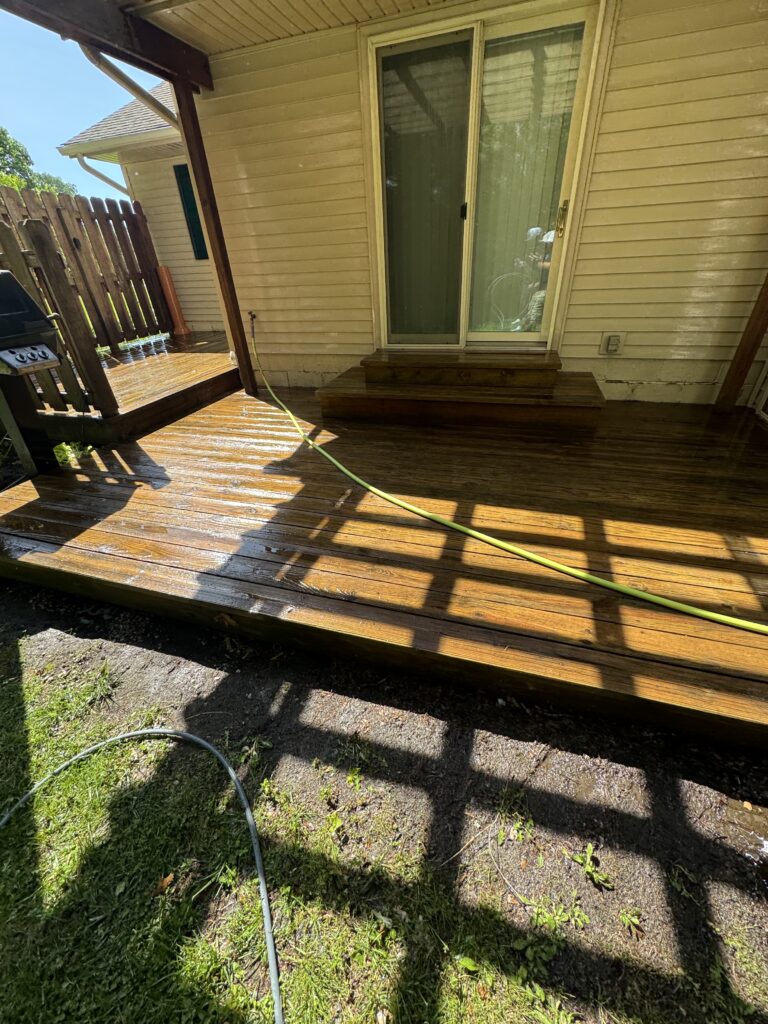 Wooden backyard deck with a sliding glass door, a green hose along wet boards, and a grill on the left under a shaded area.