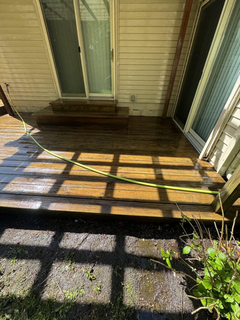 Wet wooden deck outside a house with a sliding glass door; a green garden hose crosses the boards near a small step and beige siding.