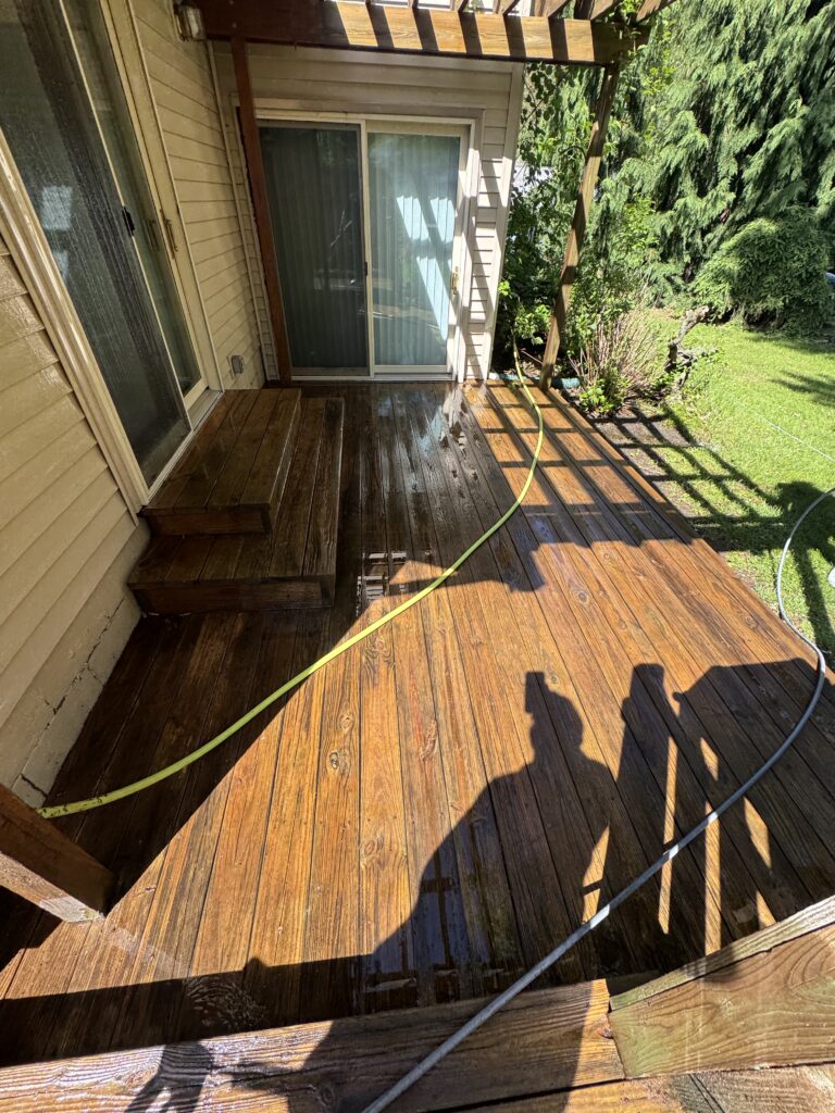 Backyard wooden deck with sliding glass door, a wet floor from a hose, and a hose lying across the boards; lawn and trees visible beyond.