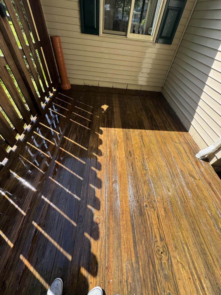 Small, sunlit wooden balcony with shadowed railing, beige siding, a window with green shutters, and an upright orange drainpipe tucked in the corner; white sneakers visible at the bottom edge.