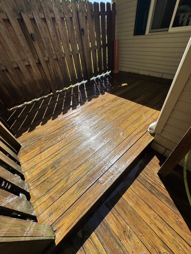 Wet wooden deck with diagonal shadows from a slatted fence, next to beige siding and a window; outdoor backyard scene.
