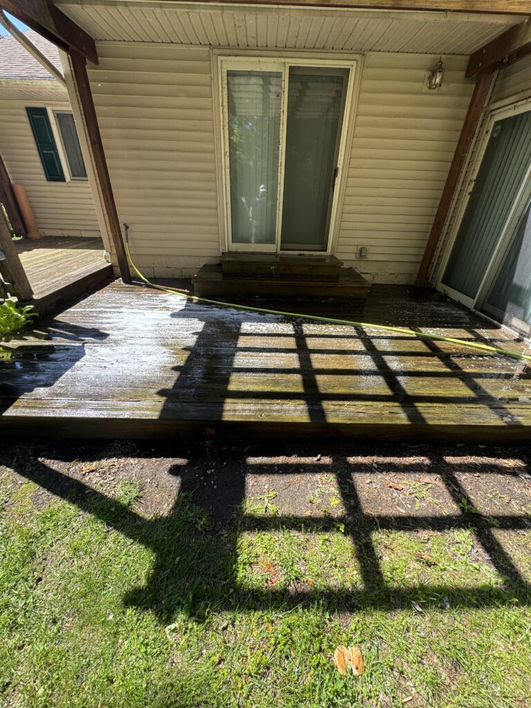 Backyard deck with a sliding glass door, wooden steps, and a yellow hose along the wet boards; railing shadows cross the deck.