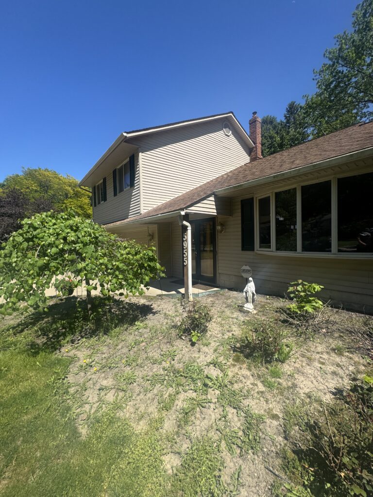 Two-story beige house with dark green shutters, brick chimney, and a sloped brown roof, set against a clear blue sky and a front yard with bushes and a statue along the path.