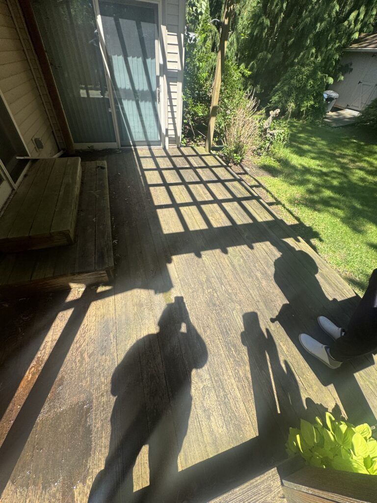 Sunlit wooden deck beside a house with a sliding glass door; long shadows from the railing create a grid pattern on the deck, a small plant in the corner and a grassy yard beyond.