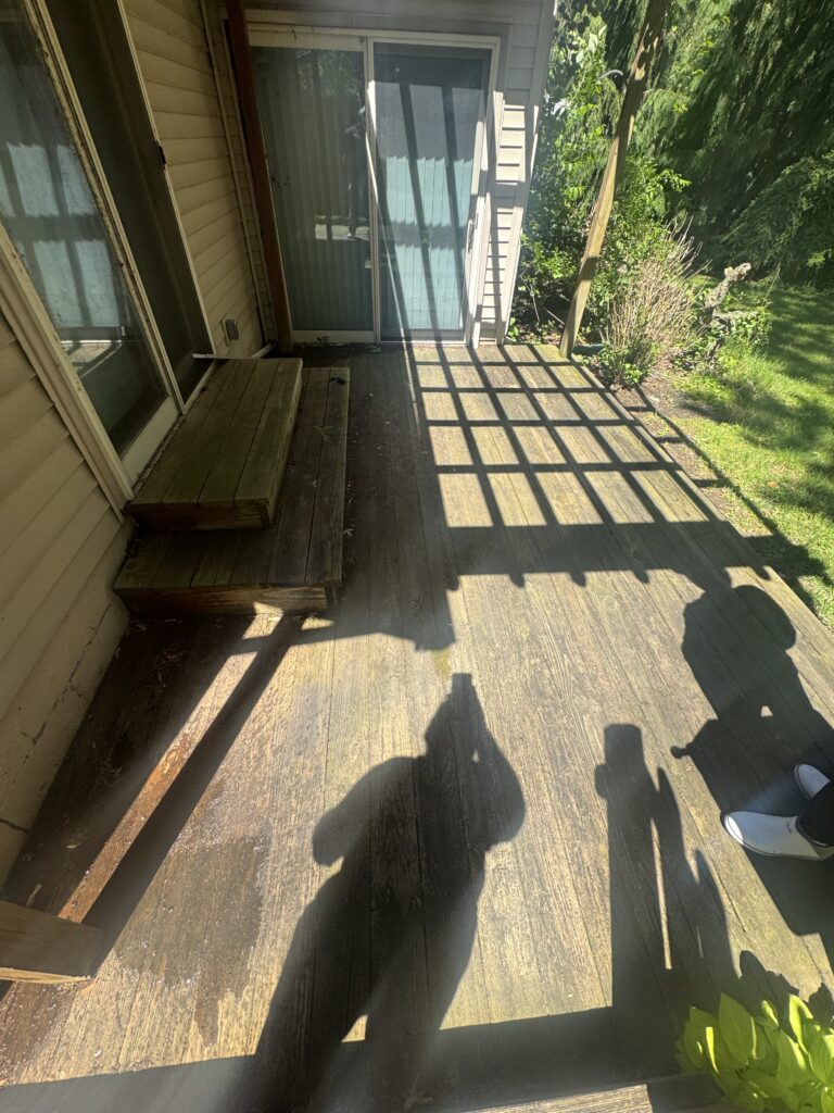 Wooden backyard deck with a sliding glass door, small steps, and sunlight casting grid-like shadows across the planks.