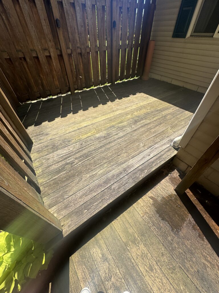 Overhead view of a small wooden deck bordered by a vertical slat privacy fence, with beige house siding on the right and a brown downspout; sunlight casts striped shadows on the deck, and a white sneaker is visible near the bottom edge.