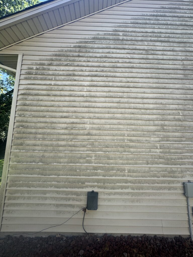 Weathered exterior wall with horizontal siding, dirt streaks, and a small black outdoor electrical box mounted mid-wall with cables trailing down to rocks at the base.