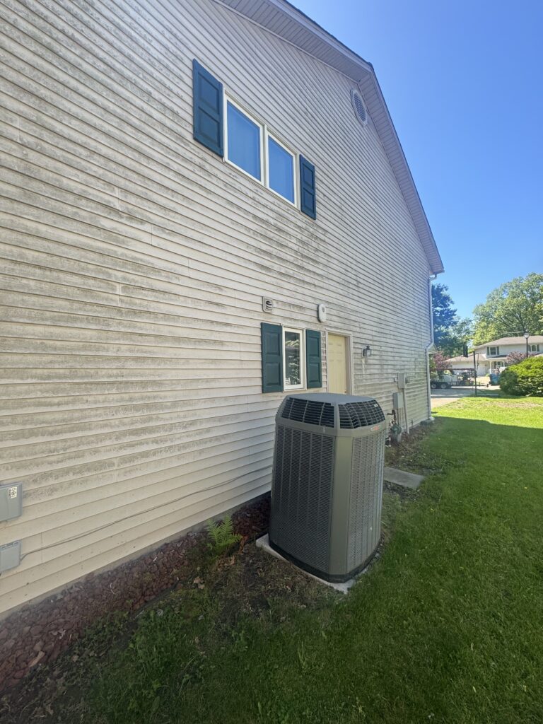 Side view of a beige siding house with blue shutters, a window, and a large outdoor air conditioner unit in the yard under a clear blue sky