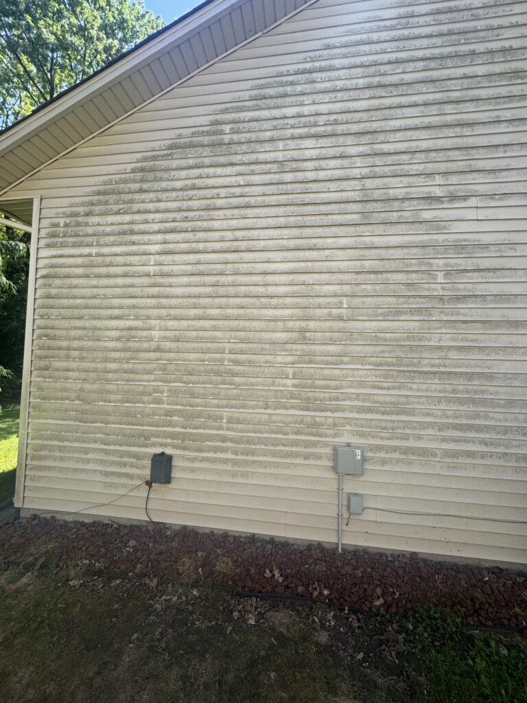 Side of a house with weathered beige vinyl siding and utility boxes mounted on the wall; dirt and rocks along the foundation, with trees visible above.