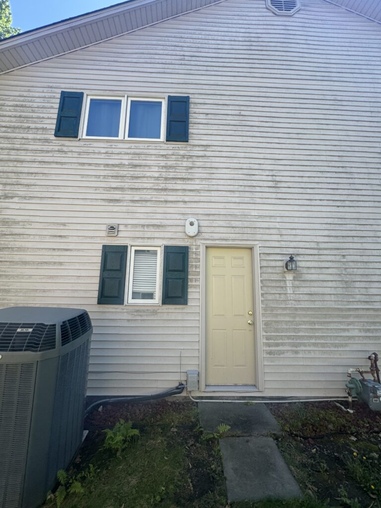Front view of a beige siding house with green shutters, a pale yellow door, and an air conditioner unit outside at left; a concrete path leads to the door.