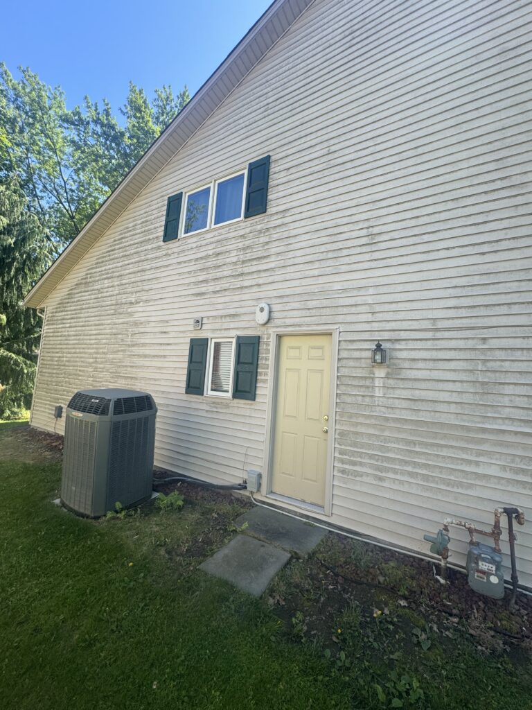 Back view of a beige house with green shutters, a yellow door, and an outdoor air conditioner unit in a grassy yard.