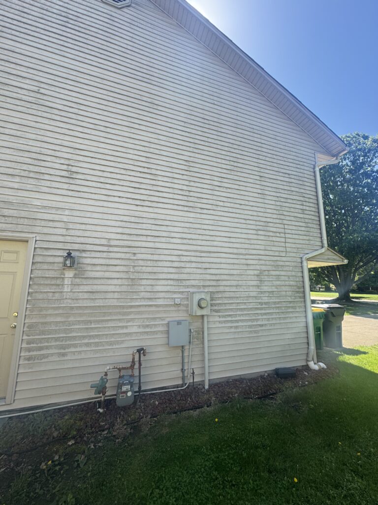 Side of a house with white siding, utility meters, and piping along the foundation; green lawn in foreground.