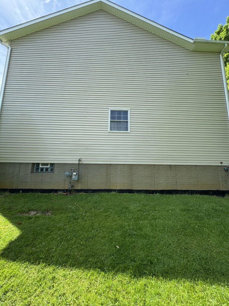 Beige vinyl-siding house wall with a small basement window, utility meters at the foundation, and a green lawn in the foreground.