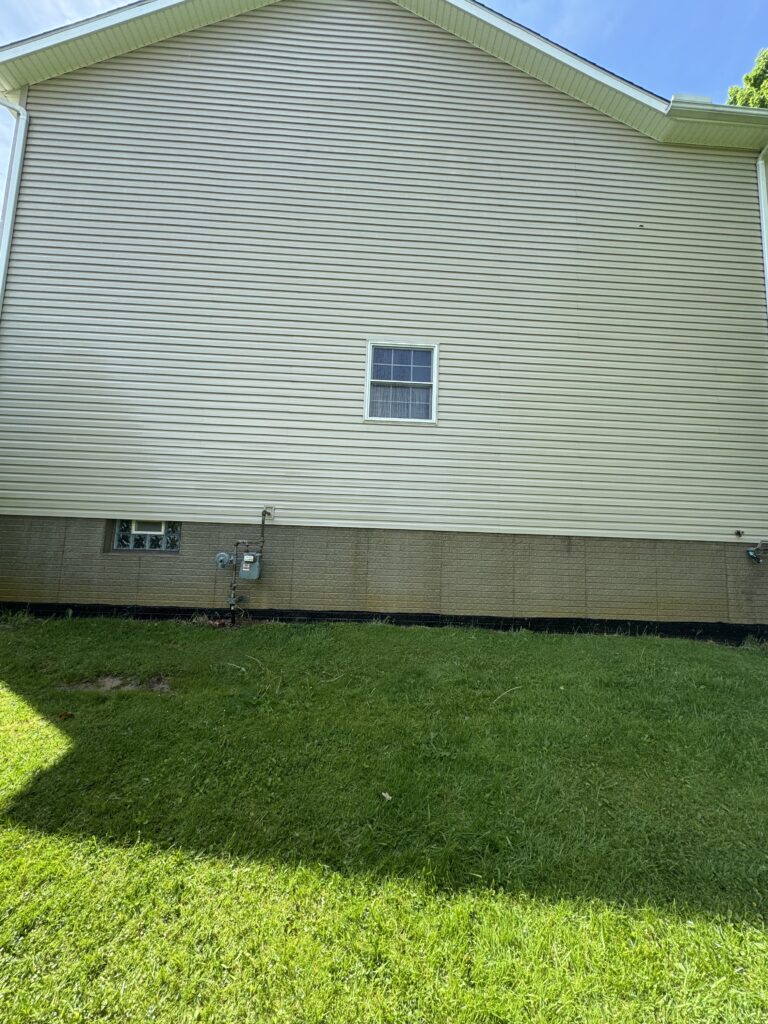 Side view of a beige vinyl-sided house with a small window and utility meters at the foundation, foreground grass visible.