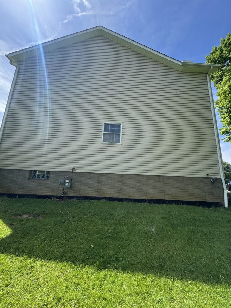 Side of a beige vinyl-sided house with a small centered window, utility meters at the foundation, and a green lawn in the foreground under a blue sky with sun glare.